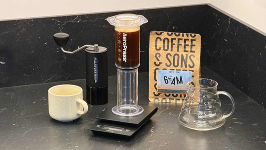 En genomskinlig AeroPress-bryggare på en digital våg, tillsammans med en handkvarn, en glaskanna och en påse Coffee & Sons-kaffe.
