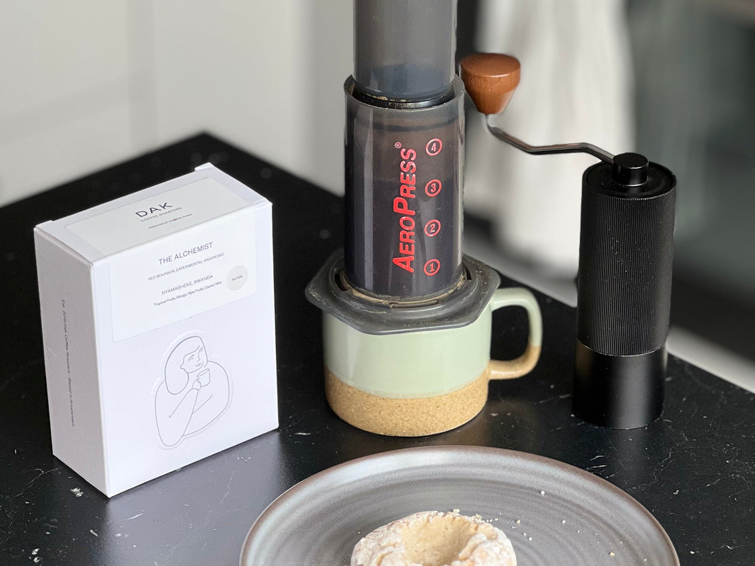 En AeroPress-kaffebryggare som vilar på en keramikmugg bredvid en låda Dak-kaffe, en manuell kvarn och ett bakverk på ett fat.