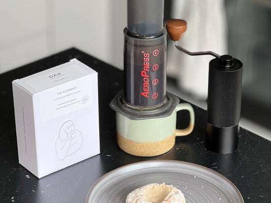 En AeroPress-kaffebryggare som vilar på en keramikmugg bredvid en låda Dak-kaffe, en manuell kvarn och ett bakverk på ett fat.