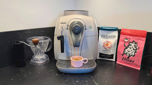 A Philips espresso machine brewing coffee into a small pink cup, displayed next to a manual grinder, a plastic Hario V60 dripper, and two bags of specialty coffee on a dark countertop.