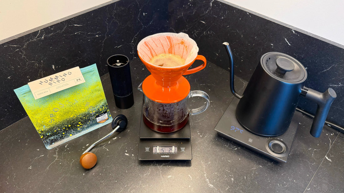 Komplett set för hemma-bryggning med en orange V60-dripper, svanhalskanna, kaffepåse och manuell kvarn på en mörk bänk.