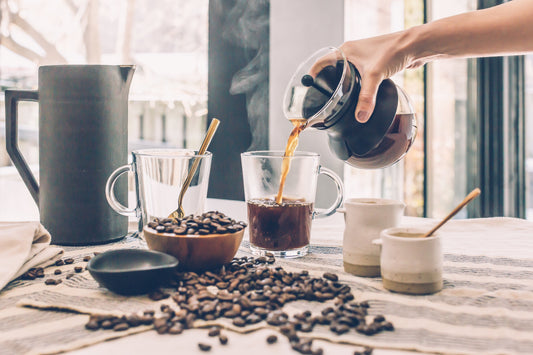 A person pouring fresh black coffee from a glass carafe into a clear mug on a table scattered with roasted coffee beans.