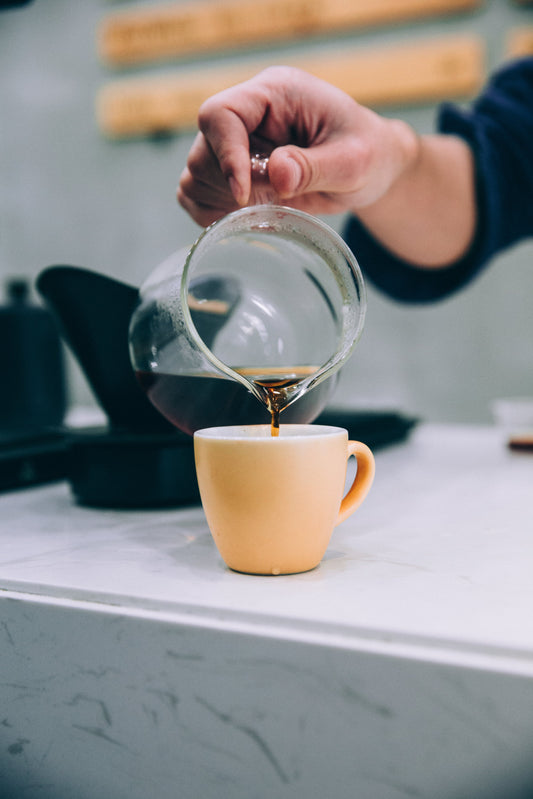 Close up of a hand pouring freshly brewed filter coffee from a glass server into a small yellow ceramic cup on a white marble counter.