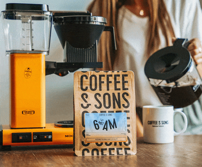 En orange Moccamaster-bryggare bredvid en påse Coffee & Sons 6AM-kaffe, med en person som häller kaffe från en kanna i en märkt vit mugg.