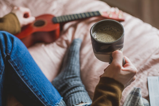 A cozy first-person perspective of a person sitting on a bed, holding a ceramic mug of coffee next to a red ukulele.