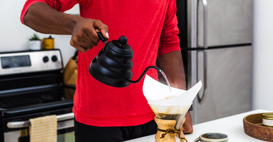 Person in a red shirt pouring coffee from a black kettle into a Chemex coffee maker on a kitchen counter.