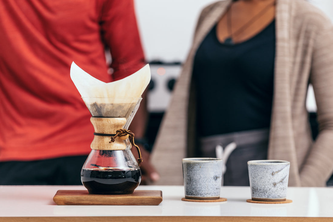 En Chemex-bryggare fylld med mörkt kaffe och två texturerade grå keramikkoppar på en vit bänk med två personer i bakgrunden.