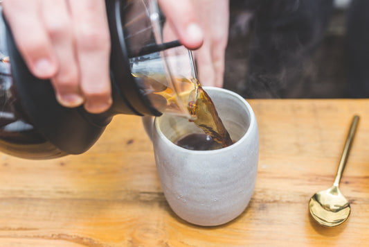 Närbild av en person som häller nybryggt kaffe från en glaskanna i en texturerad grå keramikmugg.