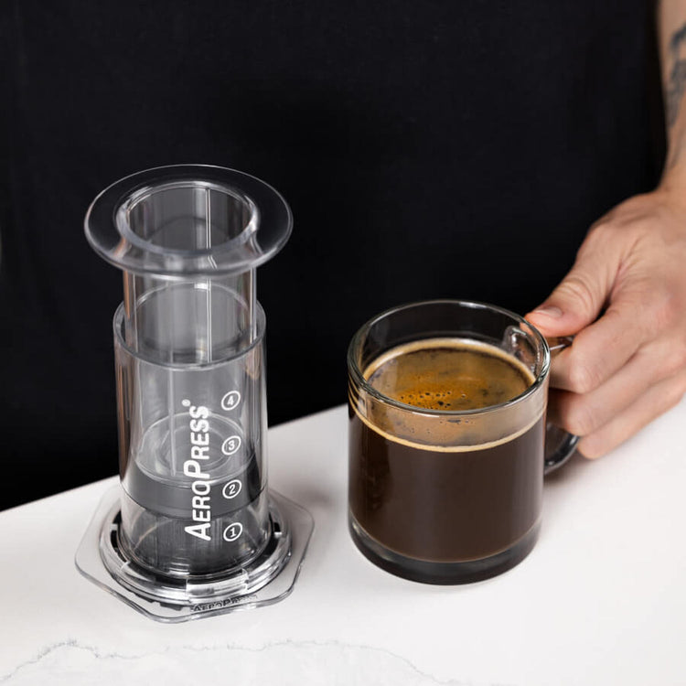 AeroPress Clear coffee maker next to a glass mug of fresh coffee.