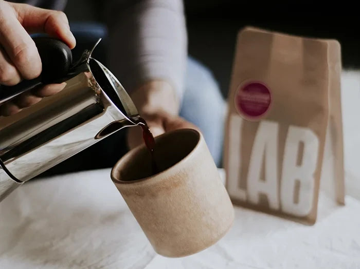 Person pouring coffee from a press into a brown paper cup with a coffee bag labeled 'LAB' in the background.