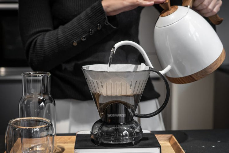 A close-up shot of a person pouring hot water from a white goose-neck kettle with wood accents into a grey Clever Dripper sitting on a coffee scale. A glass carafe and cup are on the left.