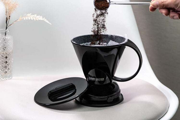 A hand using a small scoop to pour ground coffee into a paper filter lined black Clever Dripper on a white surface. The dripper lid is resting to the left, and a decorative vase with dried flowers is in the background.