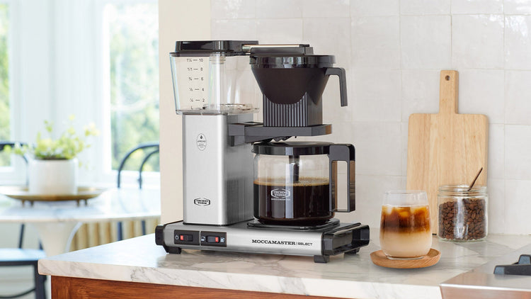 A silver Technivorm Moccamaster Select coffee maker on a marble kitchen counter next to a glass of iced coffee and a jar of coffee beans.