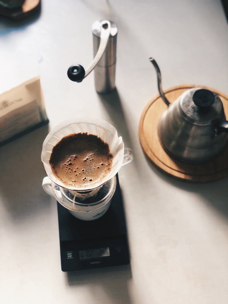 A flat lay of a pour-over coffee brewing setup, including a gooseneck kettle, a manual grinder, and coffee blooming in a dripper on a digital scale.