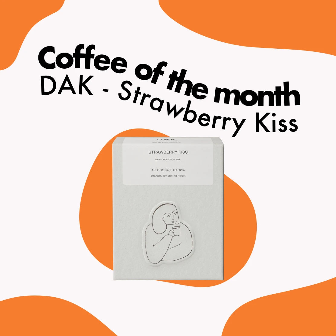 Kvadratisk banner för Månadens kaffe som visar en vit kartong DAK - Strawberry Kiss kaffe mot en vit och orange vågig bakgrund.