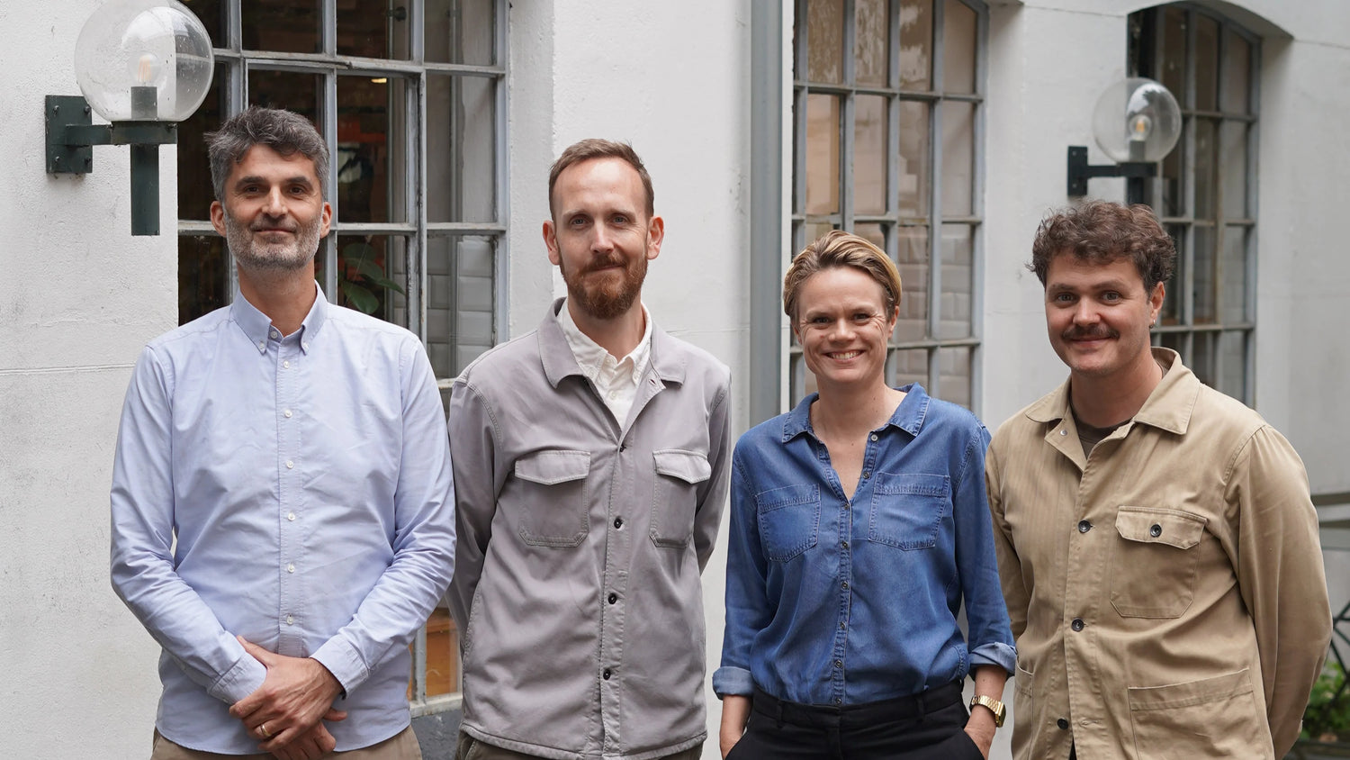 A professional portrait of four people — three men and one woman — standing together and smiling in front of a white building with industrial-style windows.