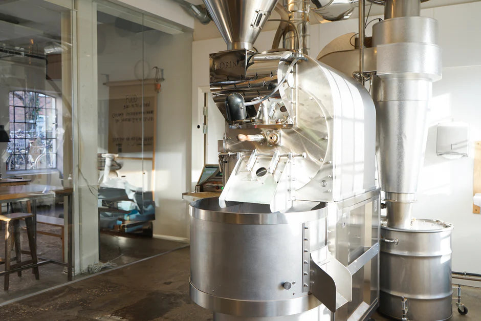 A large, industrial silver coffee roaster standing in a bright, modern roasting facility with glass walls in the background.