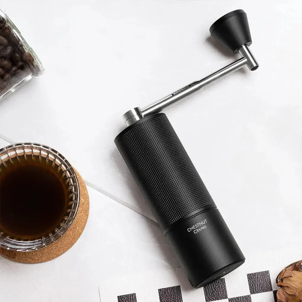 High-angle shot of a black Timemore Chestnut C3S Pro manual coffee grinder with a foldable stainless steel handle. It sits on a white surface next to a glass of brewed coffee on a cork coaster and a jar of roasted coffee beans.