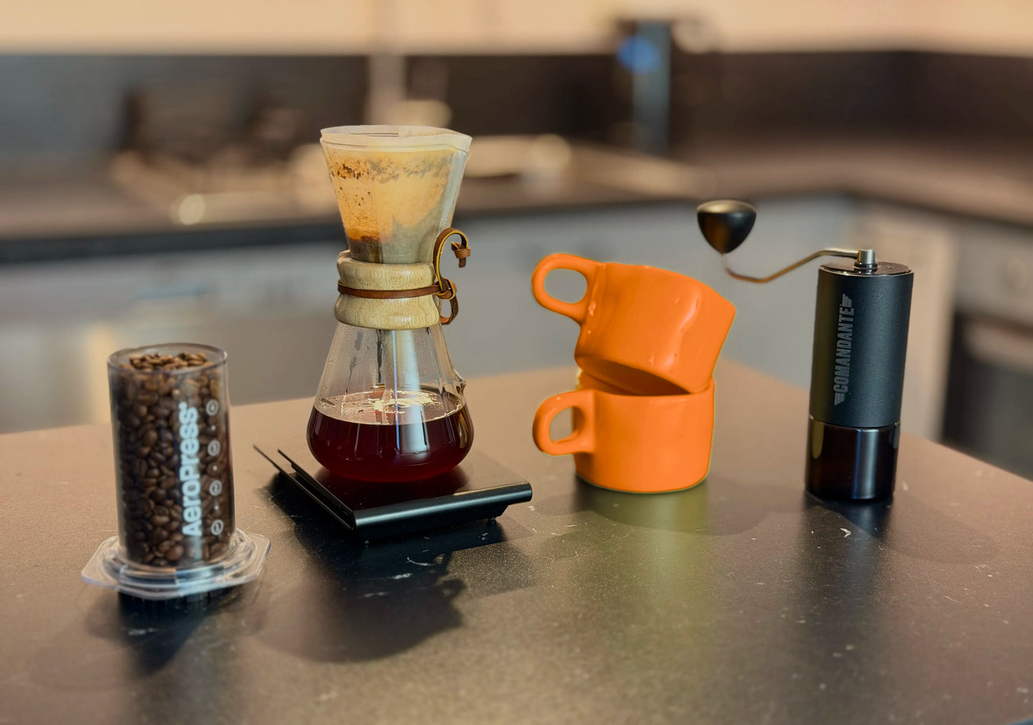 Coffee-making setup with a Chemex coffee maker, coffee beans inside Aeropress Clear, orange mugs and Comandante grinder on a kitchen counter. 