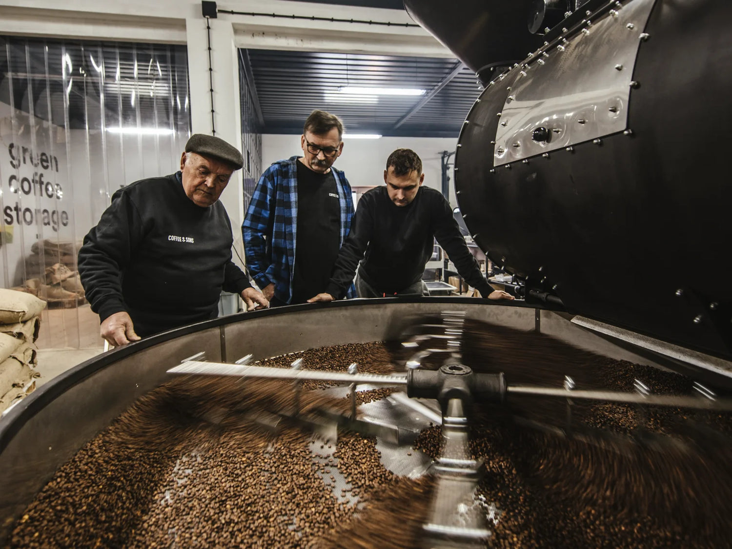 Three men inspecting a large coffee roasting machine in a warehouse setting.