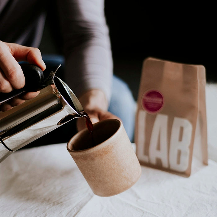Person pouring coffee from a French press into a brown mug with a coffee bag labeled 'LAB' in the background.