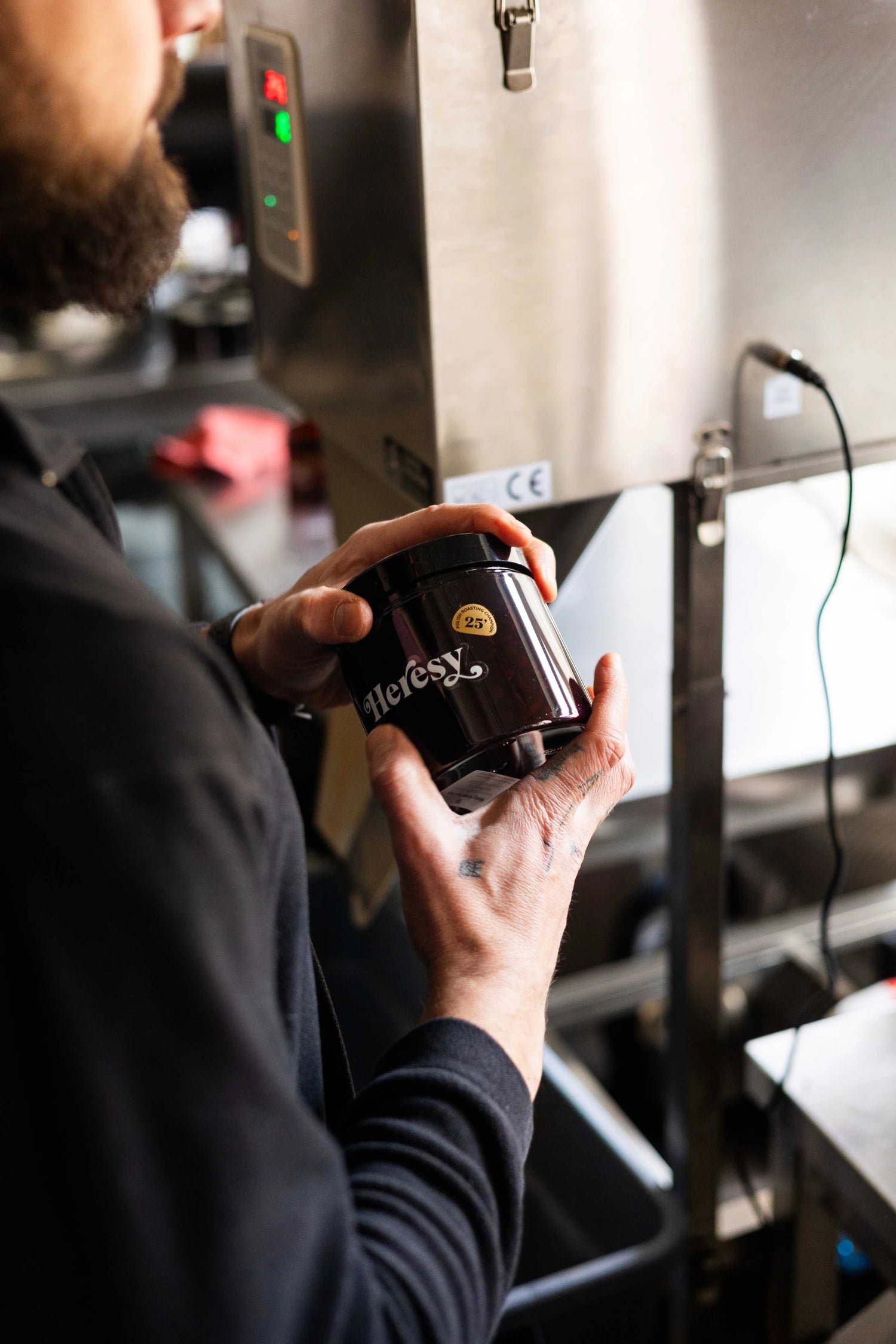 Close-up of a person holding a black Heresy brand coffee jar in front of roasting equipment.