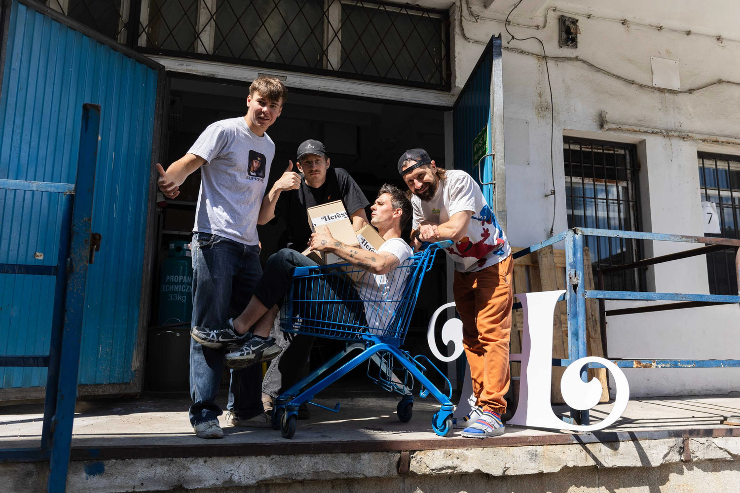 Four men posing playfully outside a warehouse, with one sitting in a blue shopping cart holding Heresy boxes.