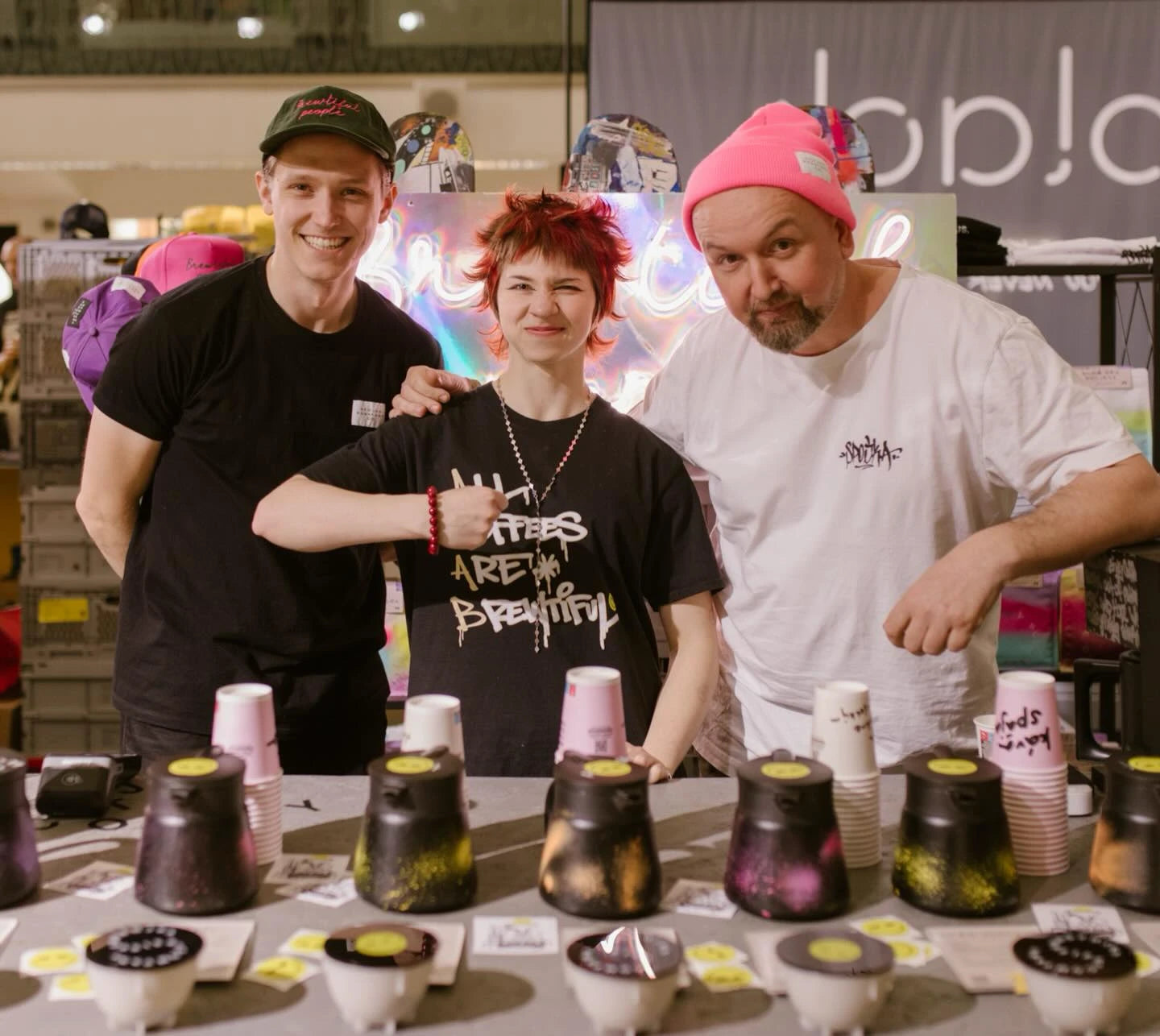 Three smiling people at a coffee booth. The central figure has red hair. In the foreground are colorful air-tight coffee containers and sample cups. A neon sign is in the background.