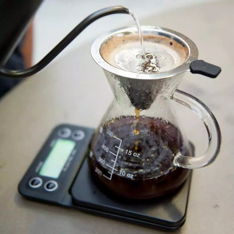 Bryggning av kaffe med gooseneck-vattenkanna och glaskaraff på en kaffevåg.