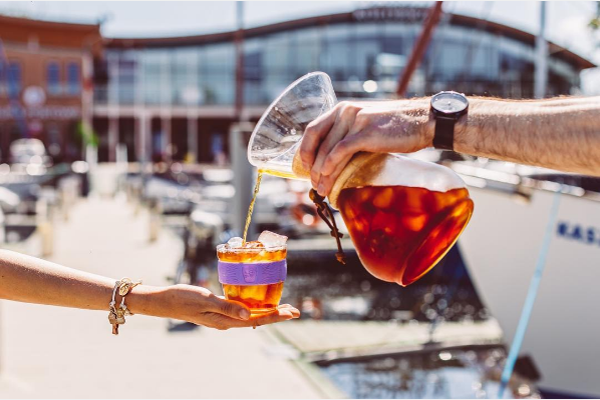 A person pouring iced coffee from a Chemex brewer into a glass with a purple sleeve, held by someone else outdoors at a sunny marina.