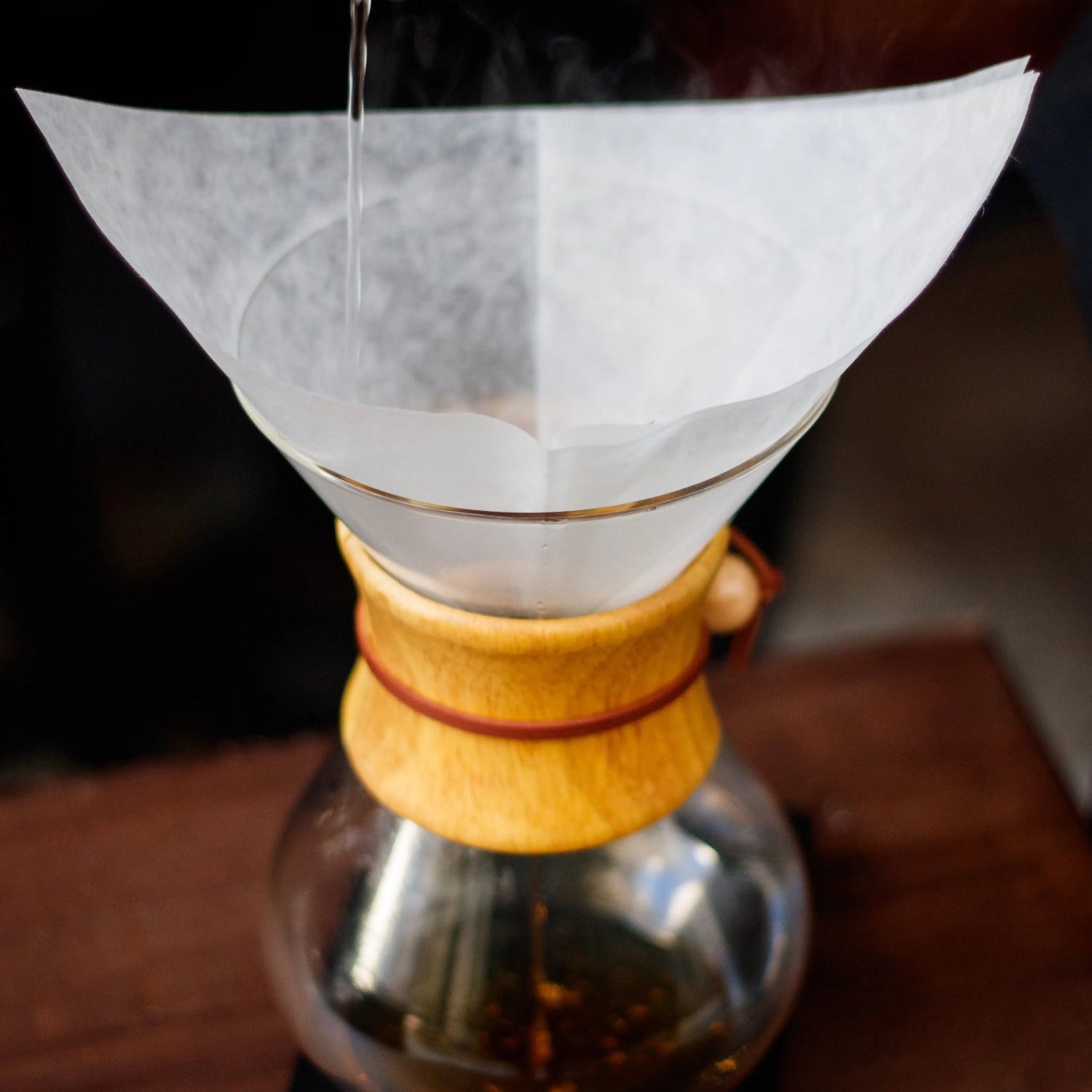 A Chemex-style pour-over coffee maker with a white paper filter cone, shown on a wooden table with hot water being poured from a metal kettle.