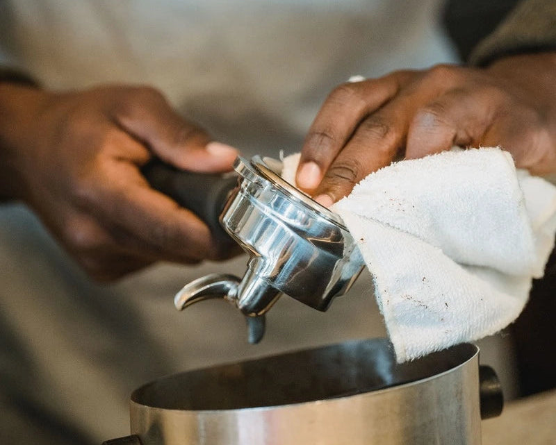 A close-up of a person's hands holding a stainless steel portafilter with a white cloth while pouring coffee into a metal pot.