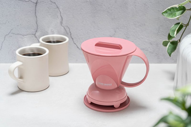A stylish product photo of a pink Clever Dripper, with its lid on and base attached, placed on a grey surface. Two beige mugs of black coffee are in the background, along with a potted plant.