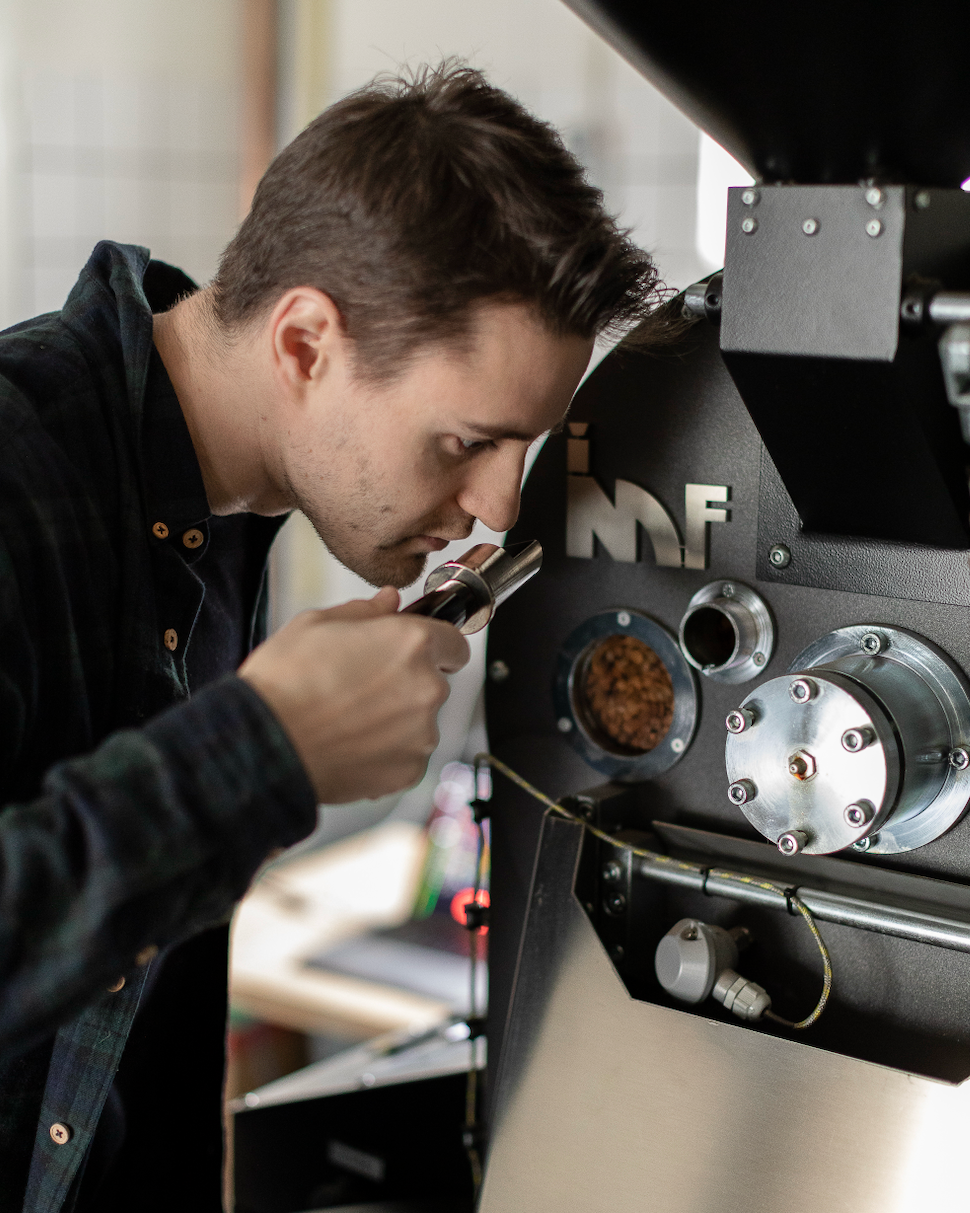A man leaning in to smell freshly roasted coffee beans from a sample trier at a large IMF roasting machine.
