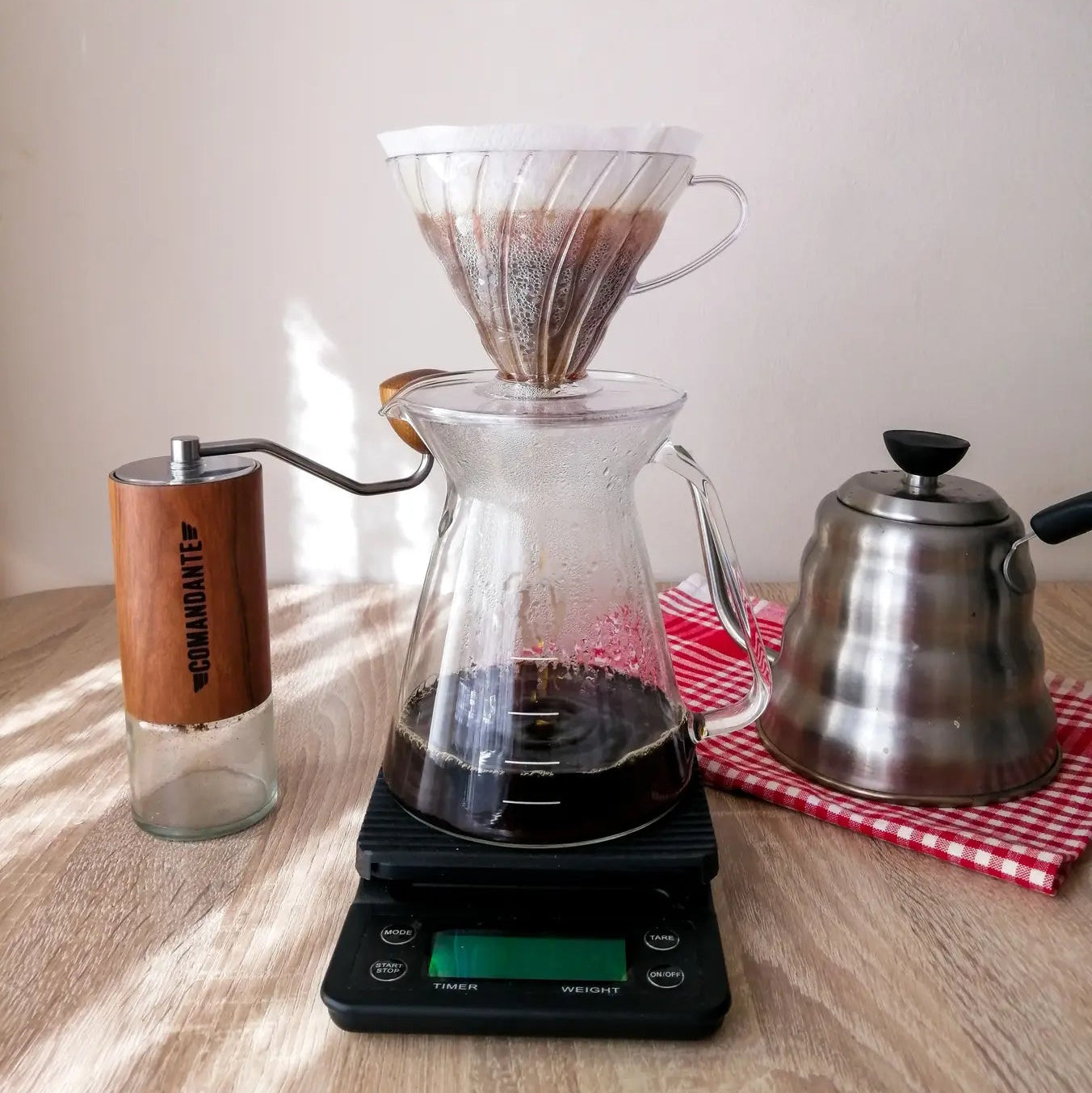 A pour-over coffee setup featuring a glass server on a digital scale with coffee dripping from a white cone filter, a manual coffee grinder and a stainless steel kettle.