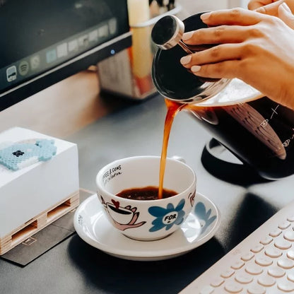 Person som häller svart kaffe från en franskpress i en vit illustrerad Coffee & Sons-kopp vid ett skrivbord