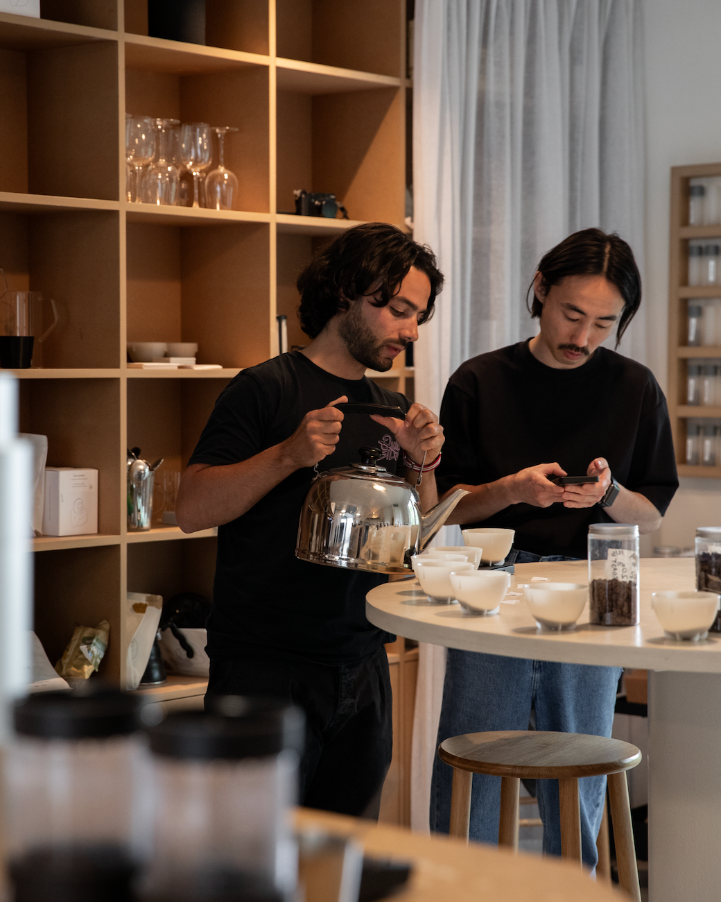 Two men in a coffee shop looking at cups where brew coffee, with shelves and a table in the background.