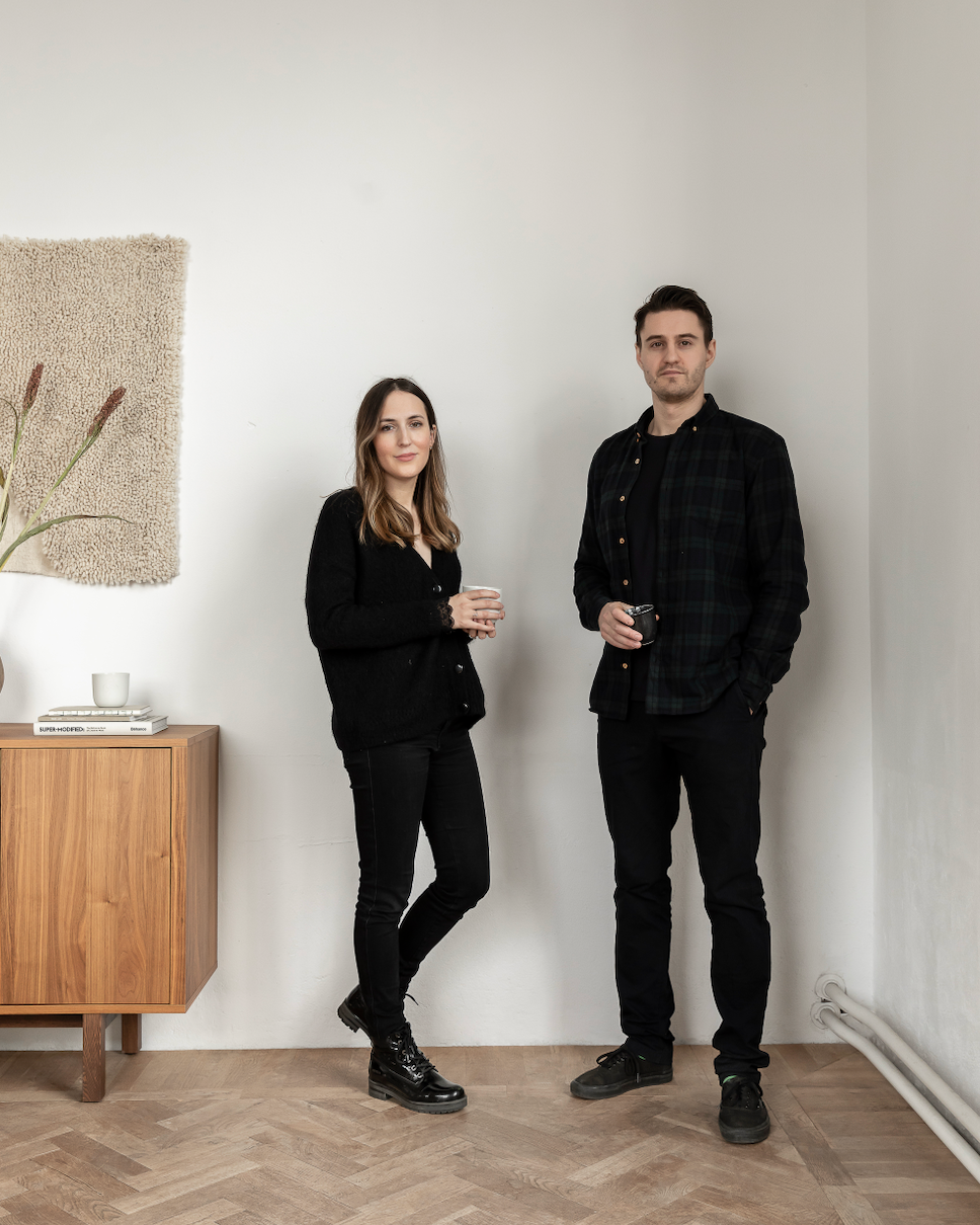 Two people standing in a room with a wooden cabinet and decorative wall art.