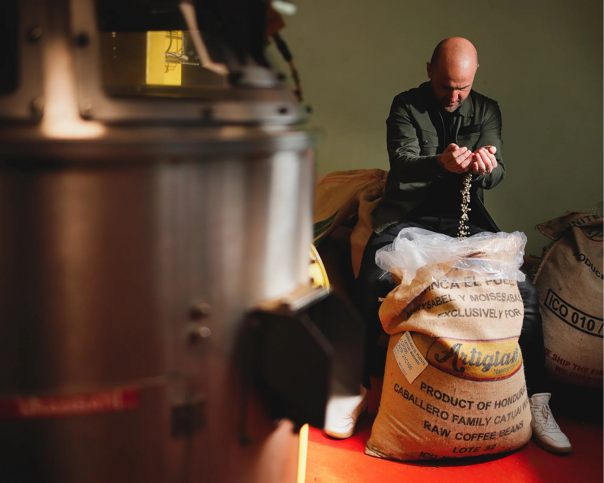 A man in a dark jacket sits on a coffee bean sack in a rustic workshop, holding a string of coffee cherries. 