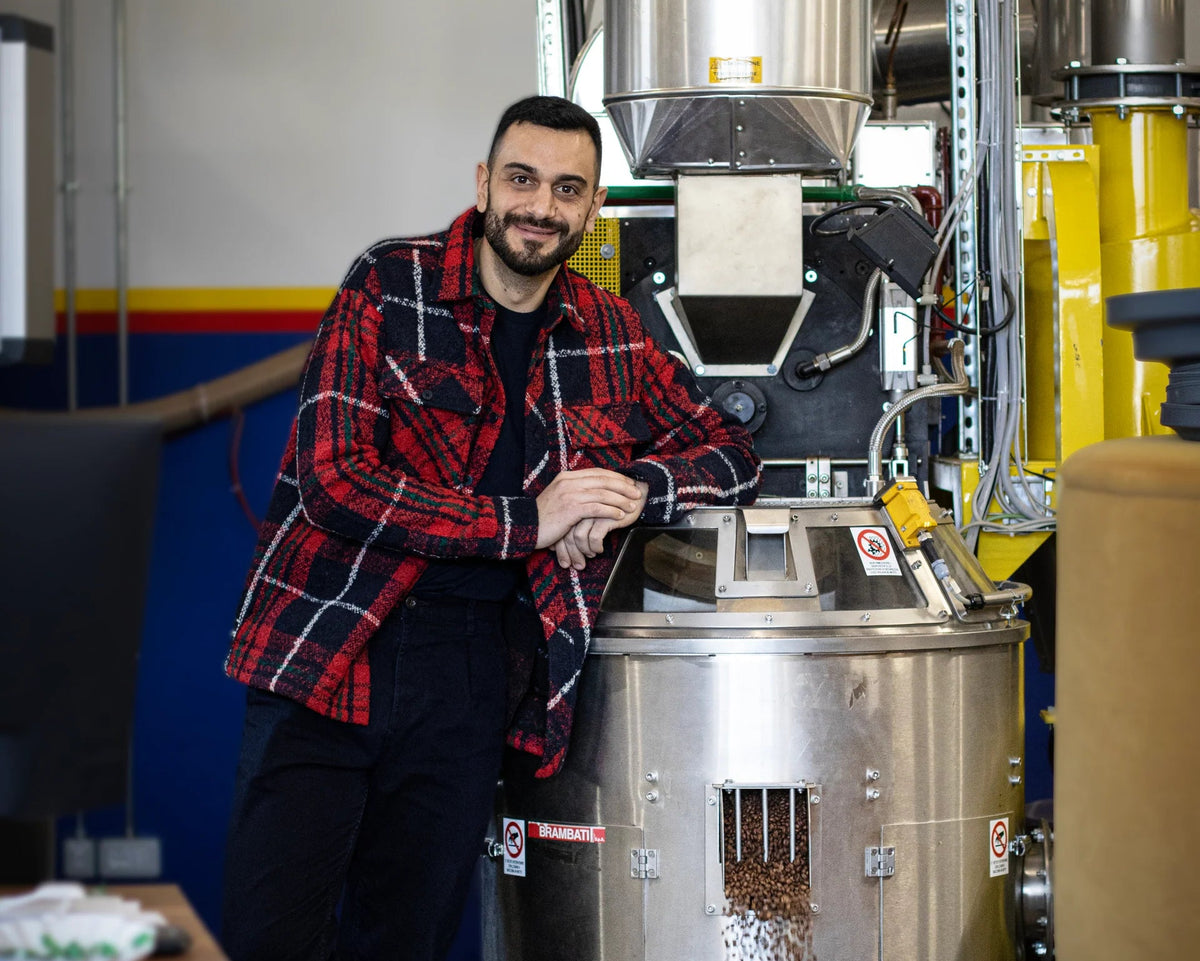 A man in a red and black plaid flannel shirt leans against a large stainless steel coffee roasting machine in a workshop setting.