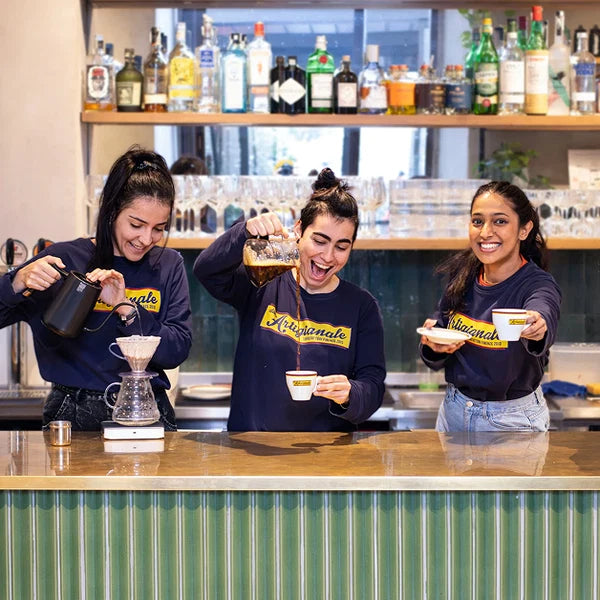 Three women smiling and laughing behind a coffee bar, wearing matching navy blue crewneck sweatshirts with a yellow graphic.