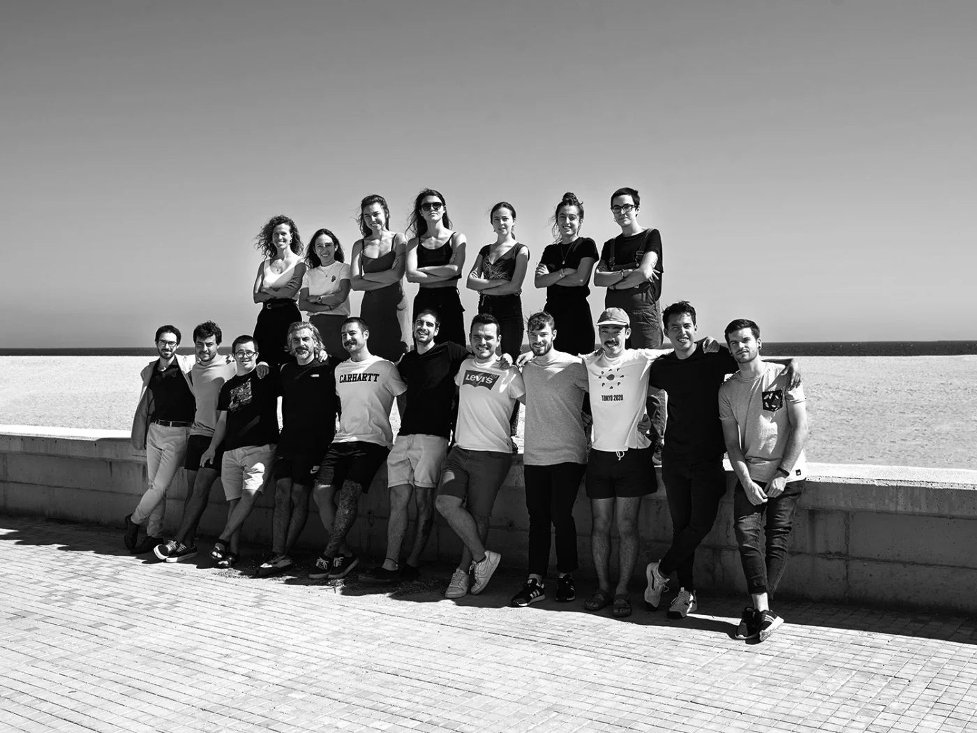 A black and white group photograph of fifteen people posing together next to a sea wall, with the ocean and clear sky in the background.