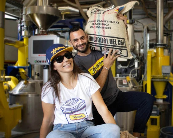 A man and a woman posing in a coffee roasting facility. The man holds a large sack of coffee beans and the woman in the foreground.
