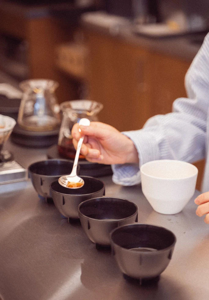 A close-up lifestyle shot of a person's hand holding a small white spoon with coffee powder, positioned over a row of black ceramic cups on a dark countertop in a cafe setting.