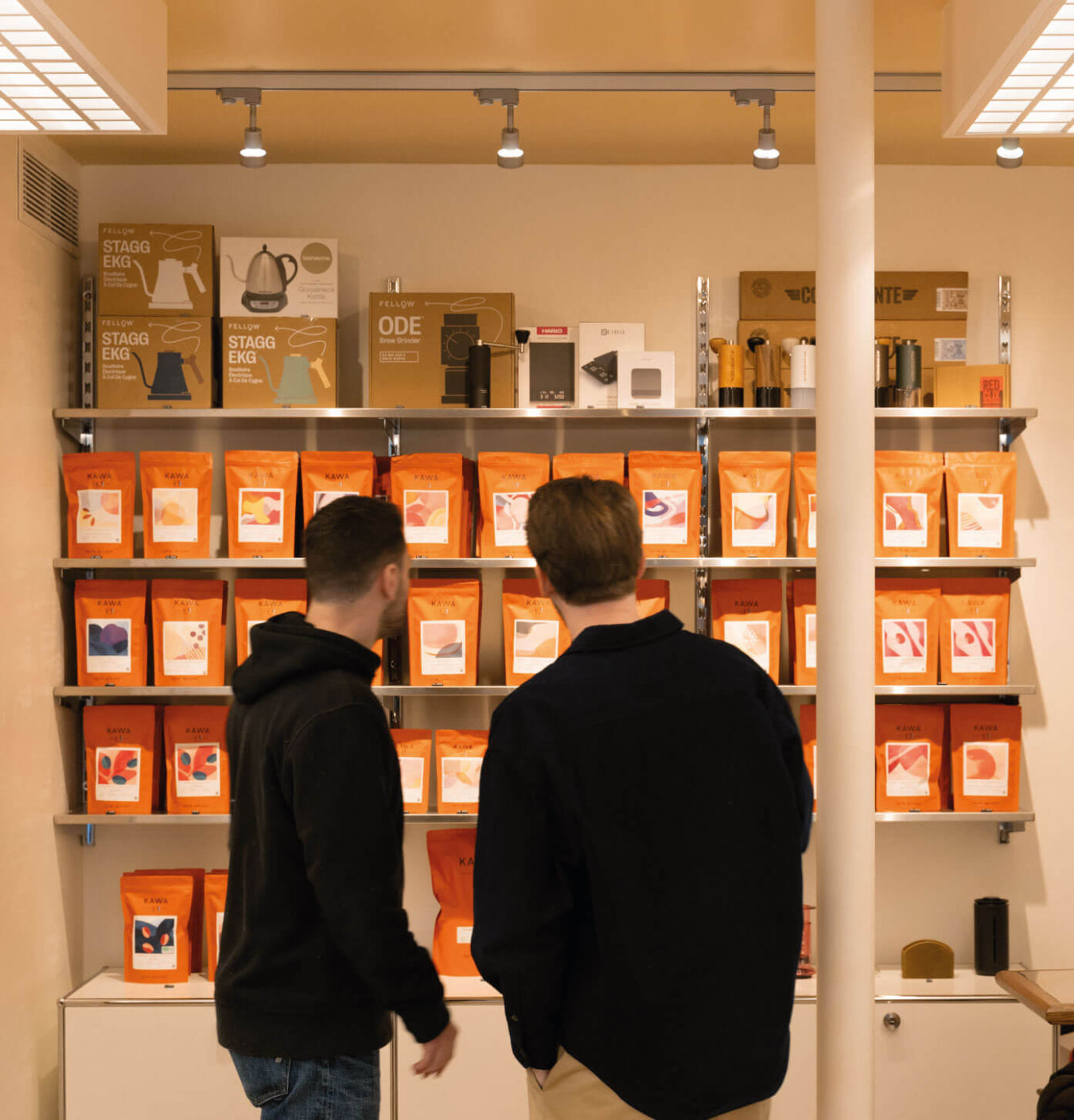 Two men viewed from behind looking at shelves filled with orange Tanat bags of coffee beans and boxes in a retail store setting.