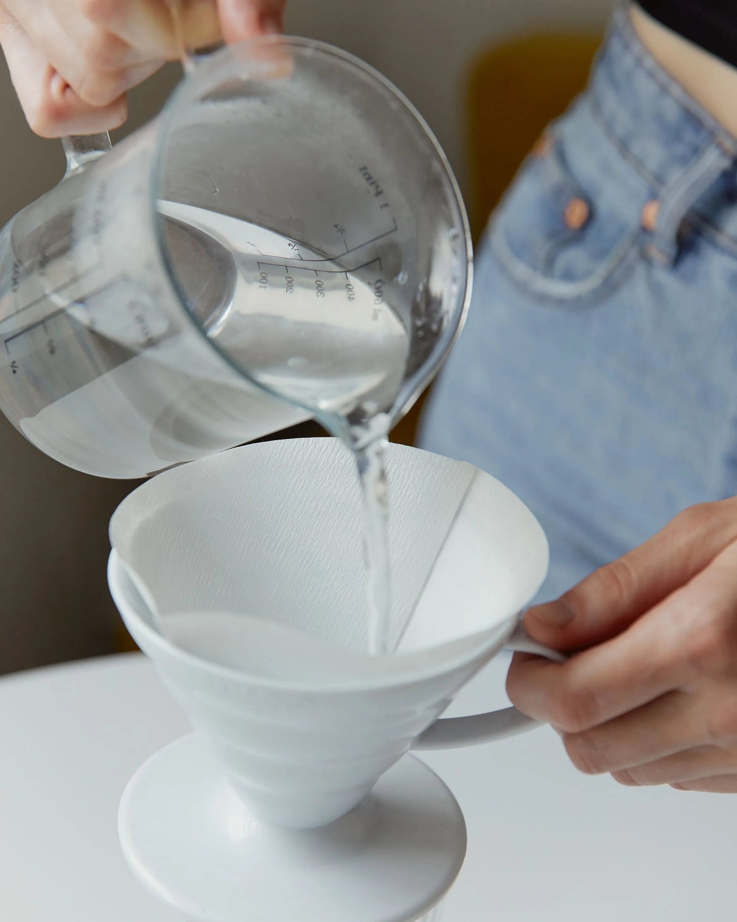 A white ceramic pour-over coffee cone with a matching saucer sits on a glass dripper, while a person pours water from a clear glass measuring pitcher into the cone. 
