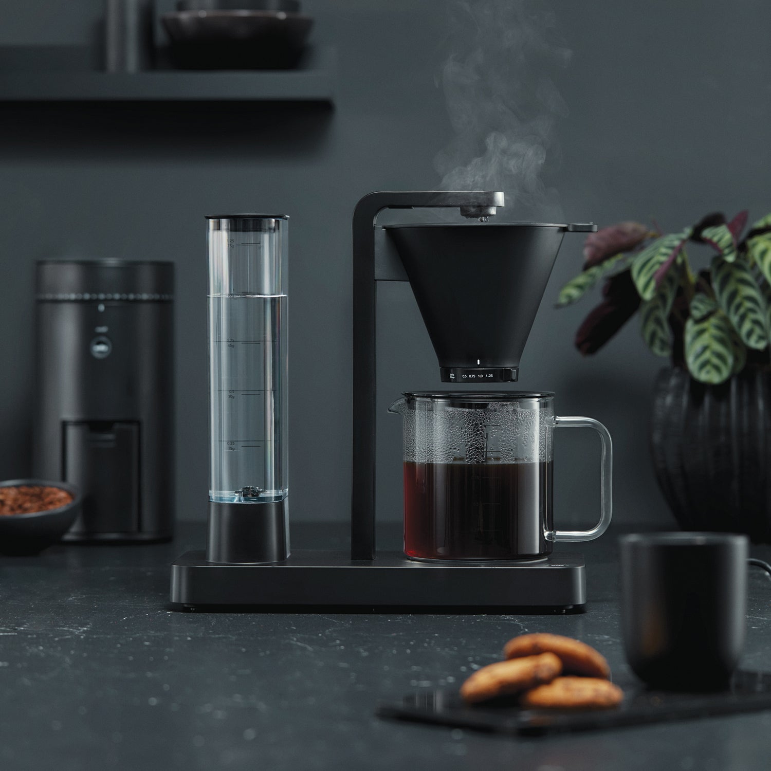 Minimalist matte black Wilfa Performance coffee brewer with a transparent water tank, showing steam rising from the filter. Set against a dark, moody background with cookies in the foreground